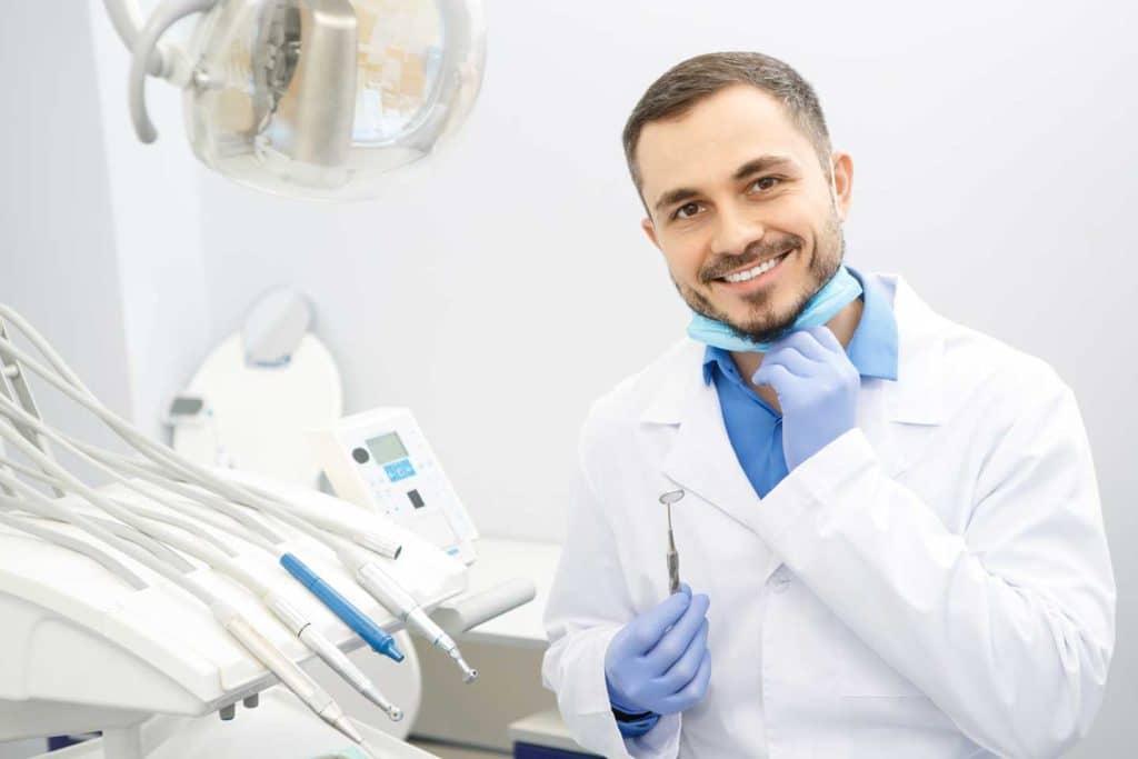 Professional male dentist smiling to the camera holding dental instruments sitting at his office