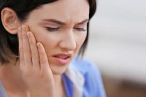 Woman in pain grimacing and holding cheek, possibly experiencing a toothache in need of a tooth-coloured filling